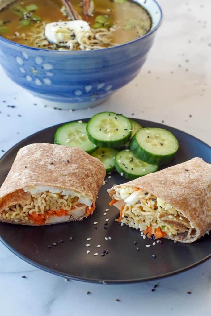 Ramen wrap on a black plate with cucumbers and ramen soup in the background.