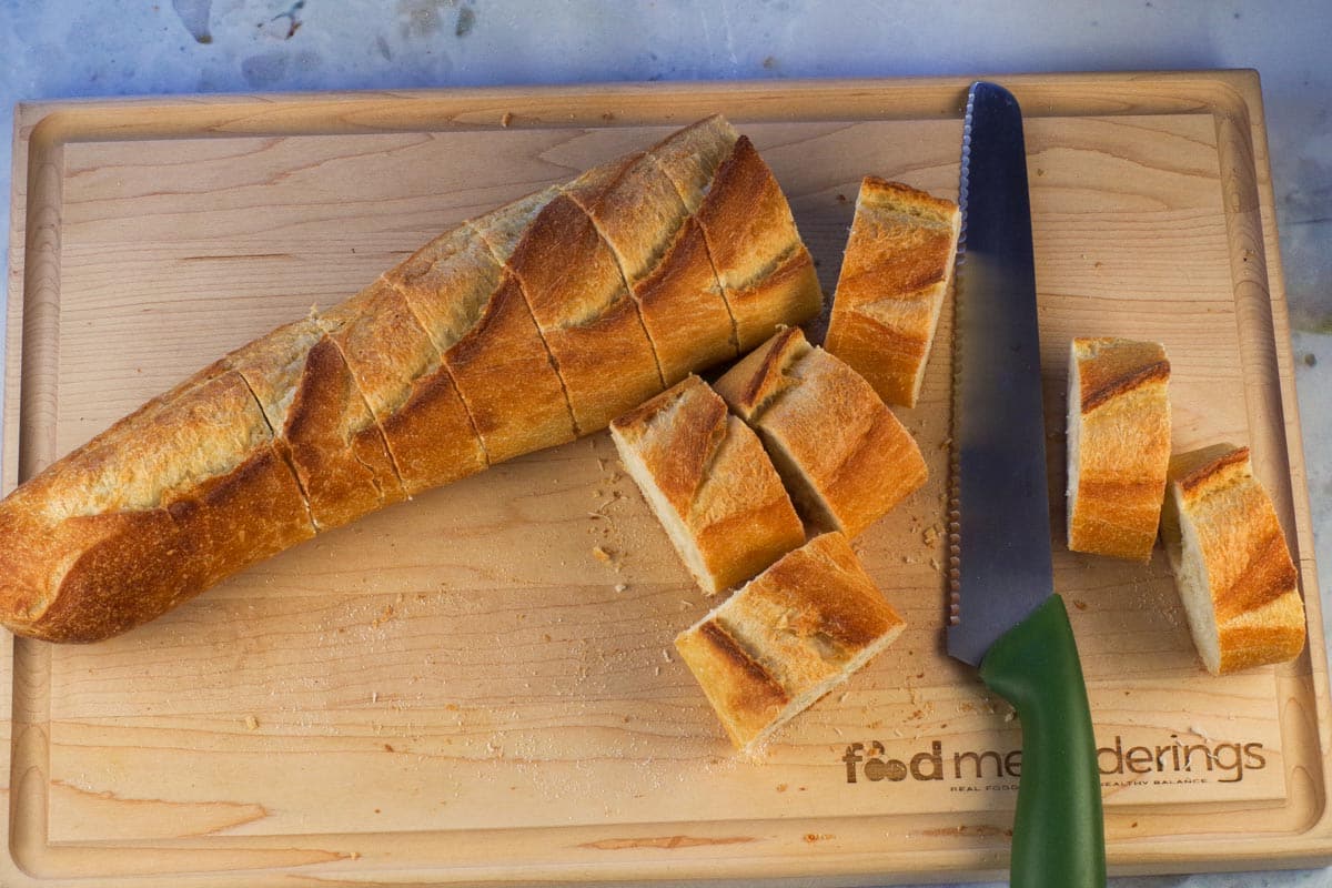 Baguette cut into 1 inch pieces on a wooden cutting  board with a bread knife.