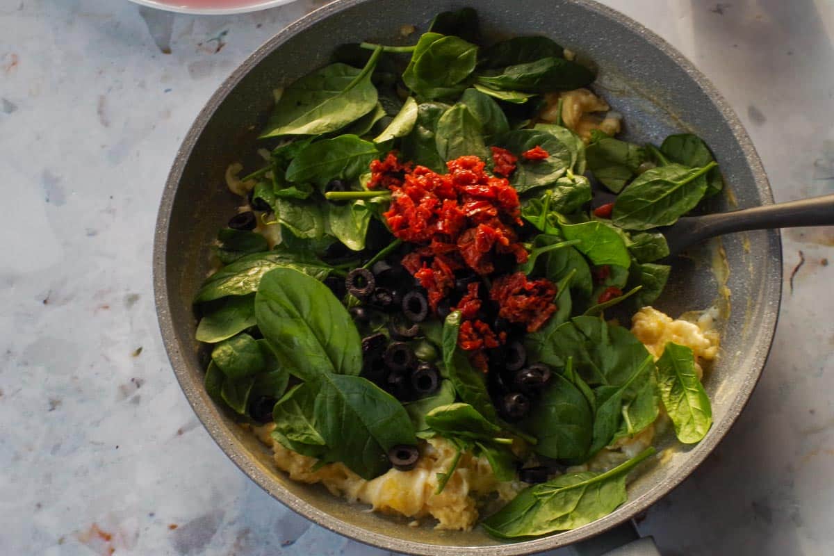 Spinach. olives and sun-dried tomatoes added to eggs in frying pan.