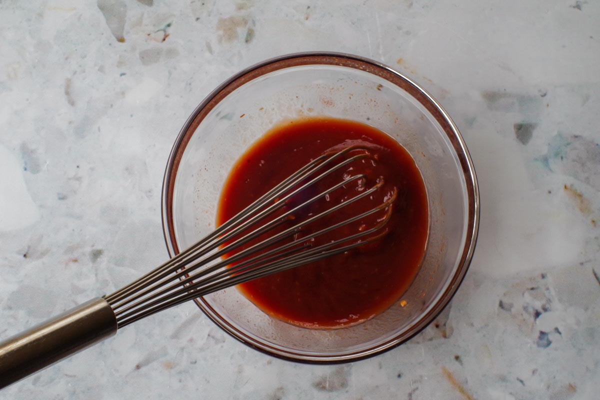 Ingredients in Barbeque sauce with ketchup and brown sugar mixed together in a glass bowl with a large metal whisk.