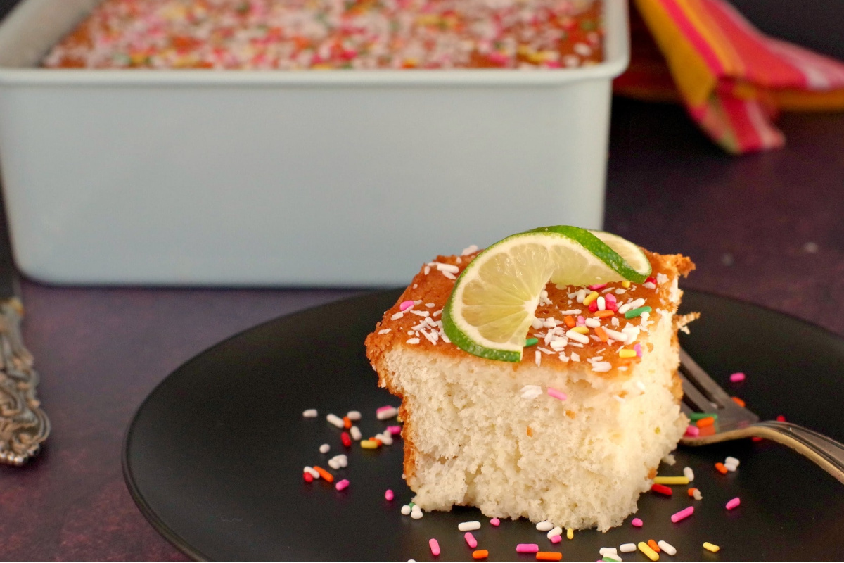 A piece of Coconut Tres Leche Cake with sprinkles and a lime twist on a black plate, with an 8x8 pan of more cake in the background. 