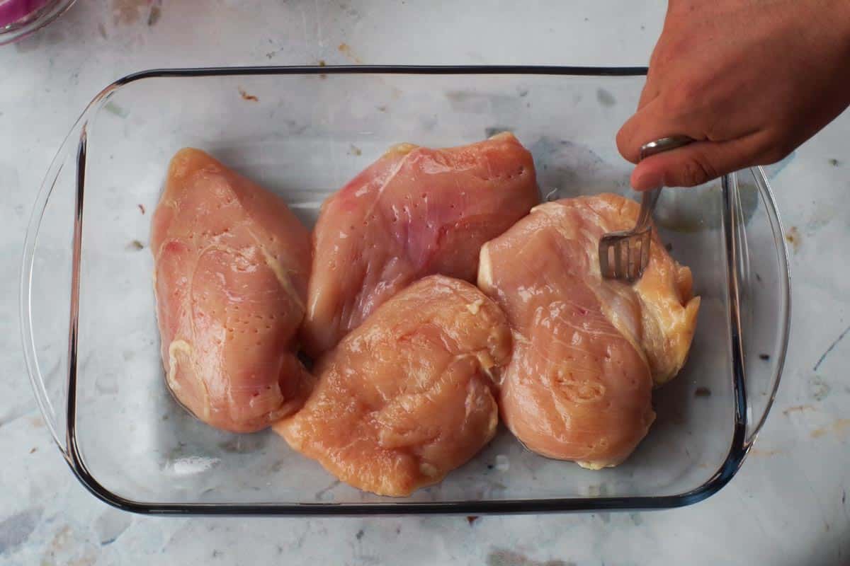 Raw chicken breasts in a glass container being scored with a fork.