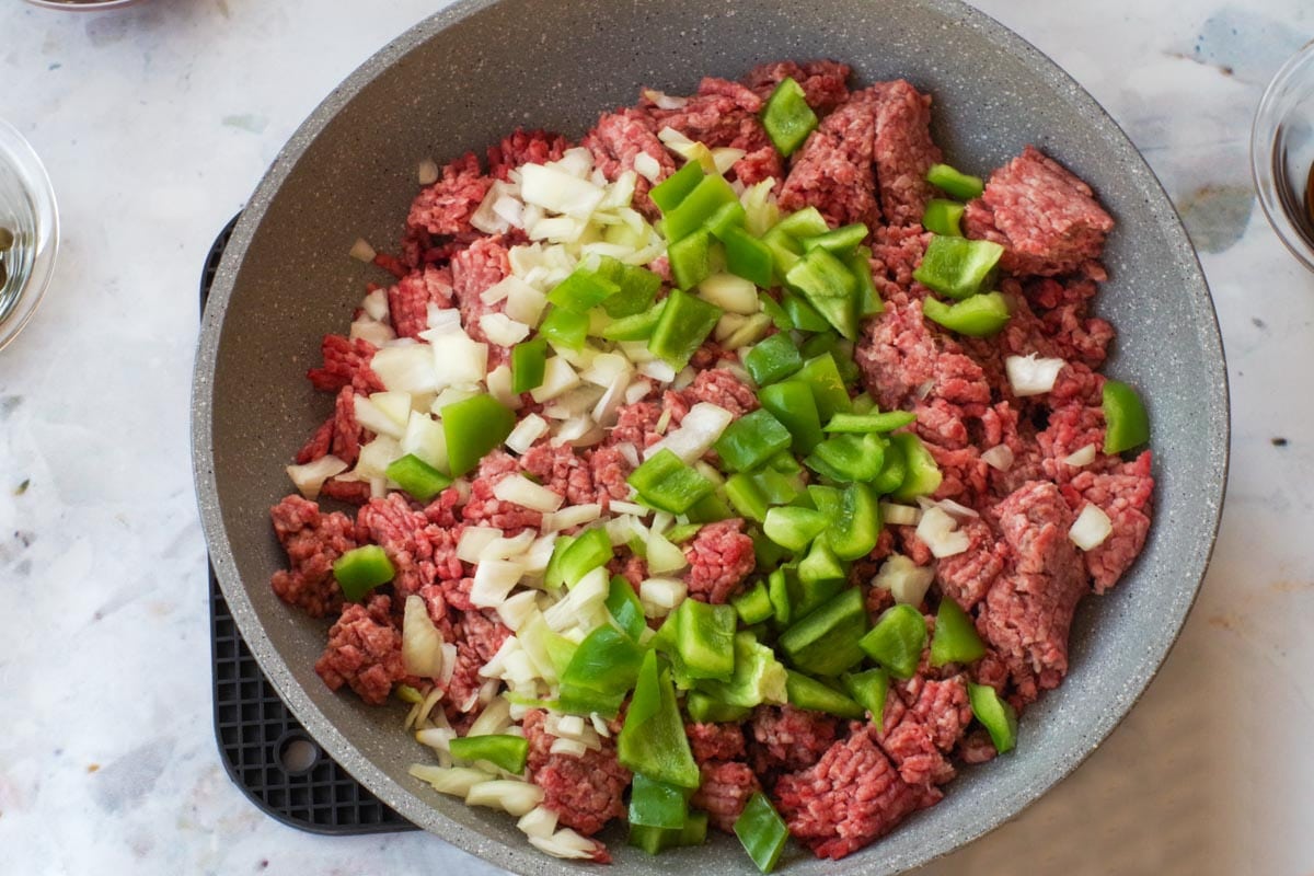 Raw ground beef in frying pan with uncooked onions and green bell peppers.