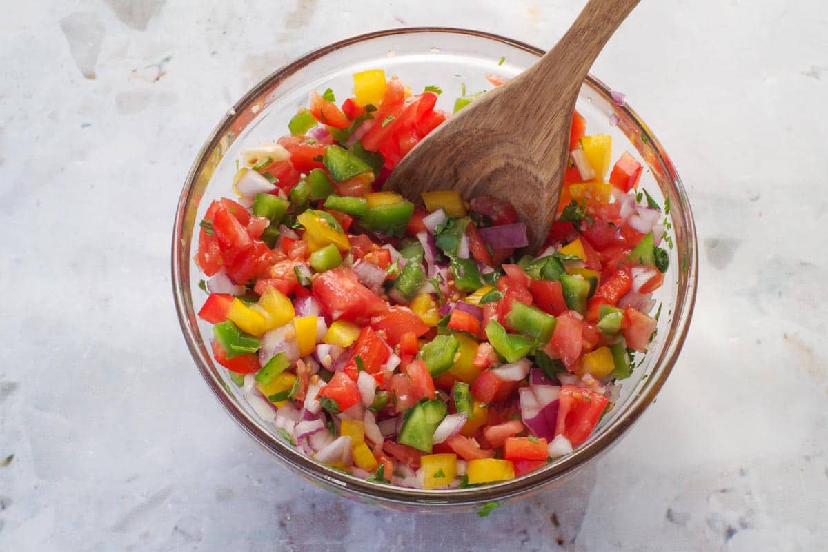 Pico de Gallo Salsa ingredients mixed together in a medium glass bowl with a wooden spoon.