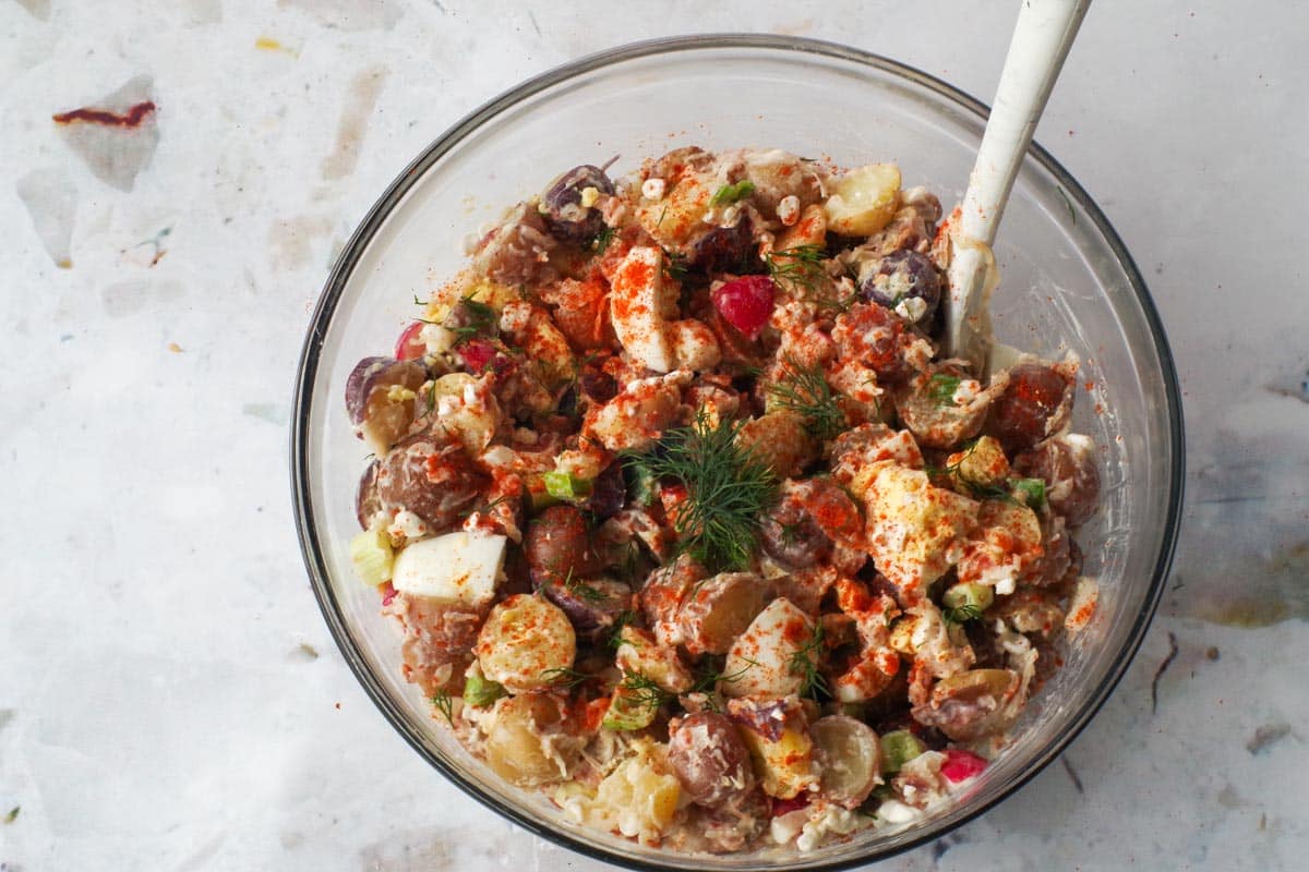 Garnishes added to potato salad in large glass bowl with spoon.