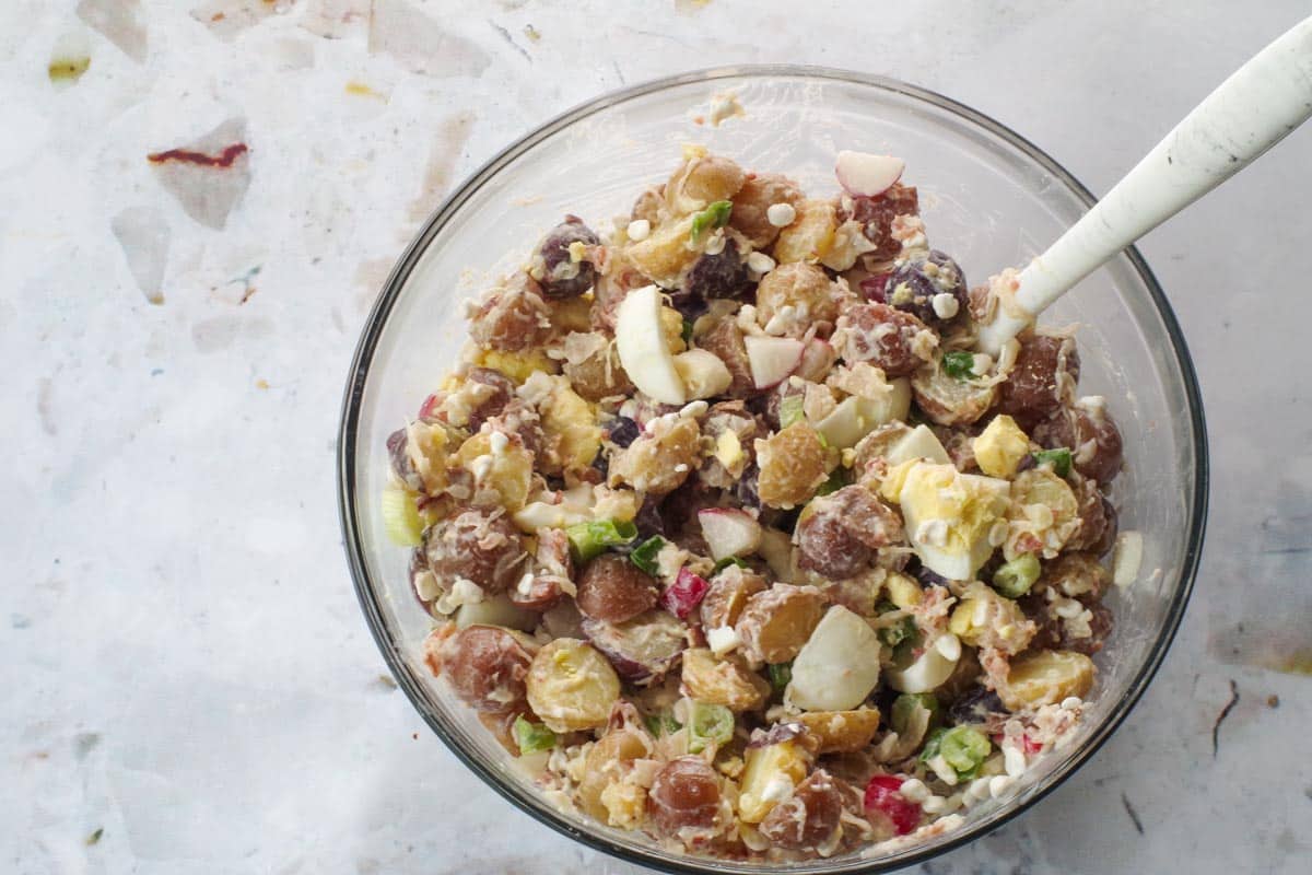 Eggs added to potato salad in large glass bowl with marble silicone spoon.