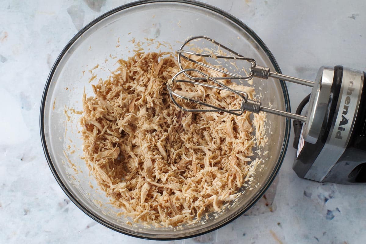 Cooked chicken breast shredded with mixer in glass bowl.