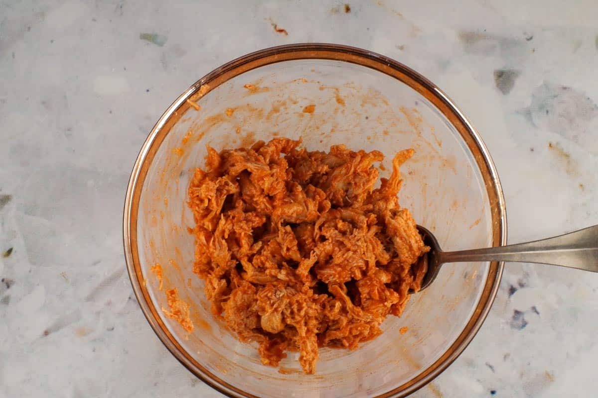 Pulled buffalo chicken in a glass mixing bowl with a spoon.