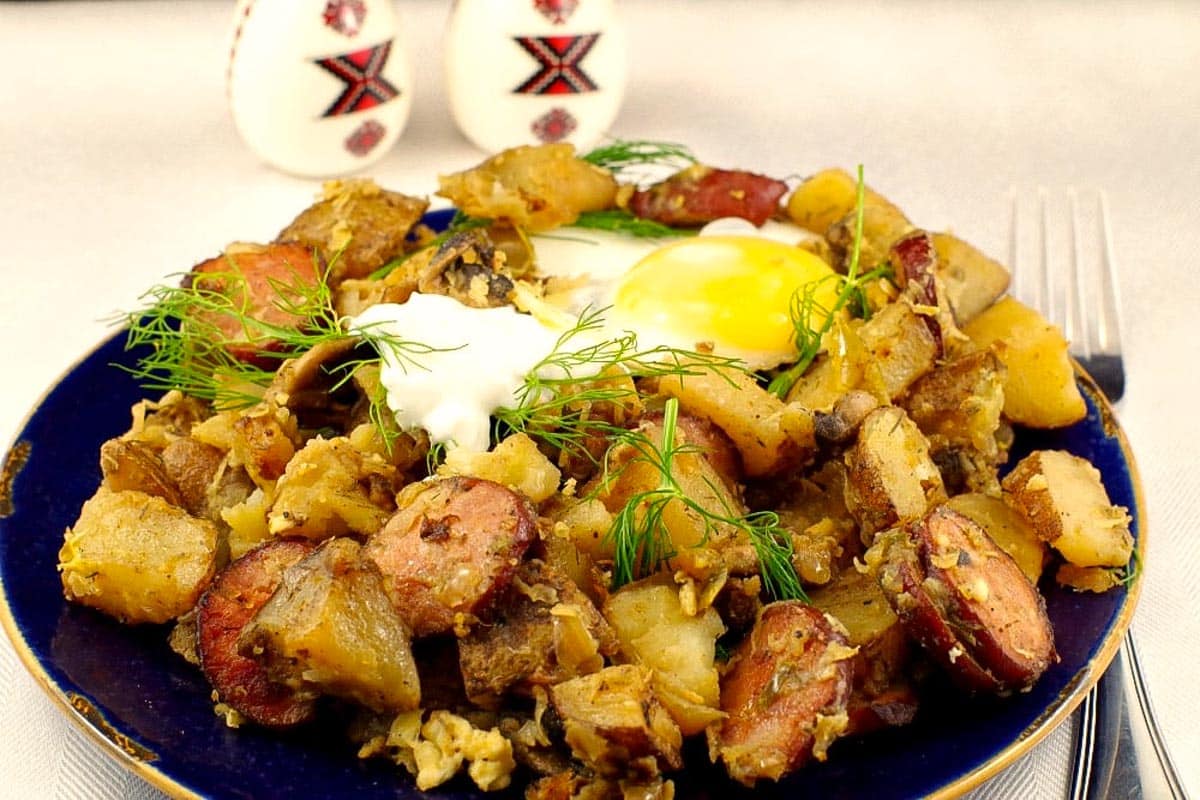 Ukrainian Breakfast Hash on a plate with a fried egg on top. and Ukrainian patterned salt and pepper shakers in the background.