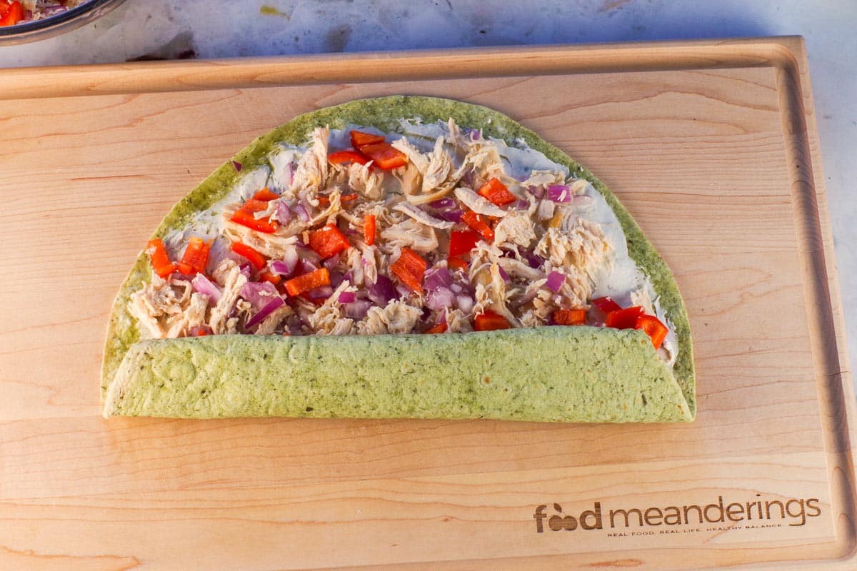 Chicken tortilla pinwheels being rolled up from bottom on wooden cutting board.