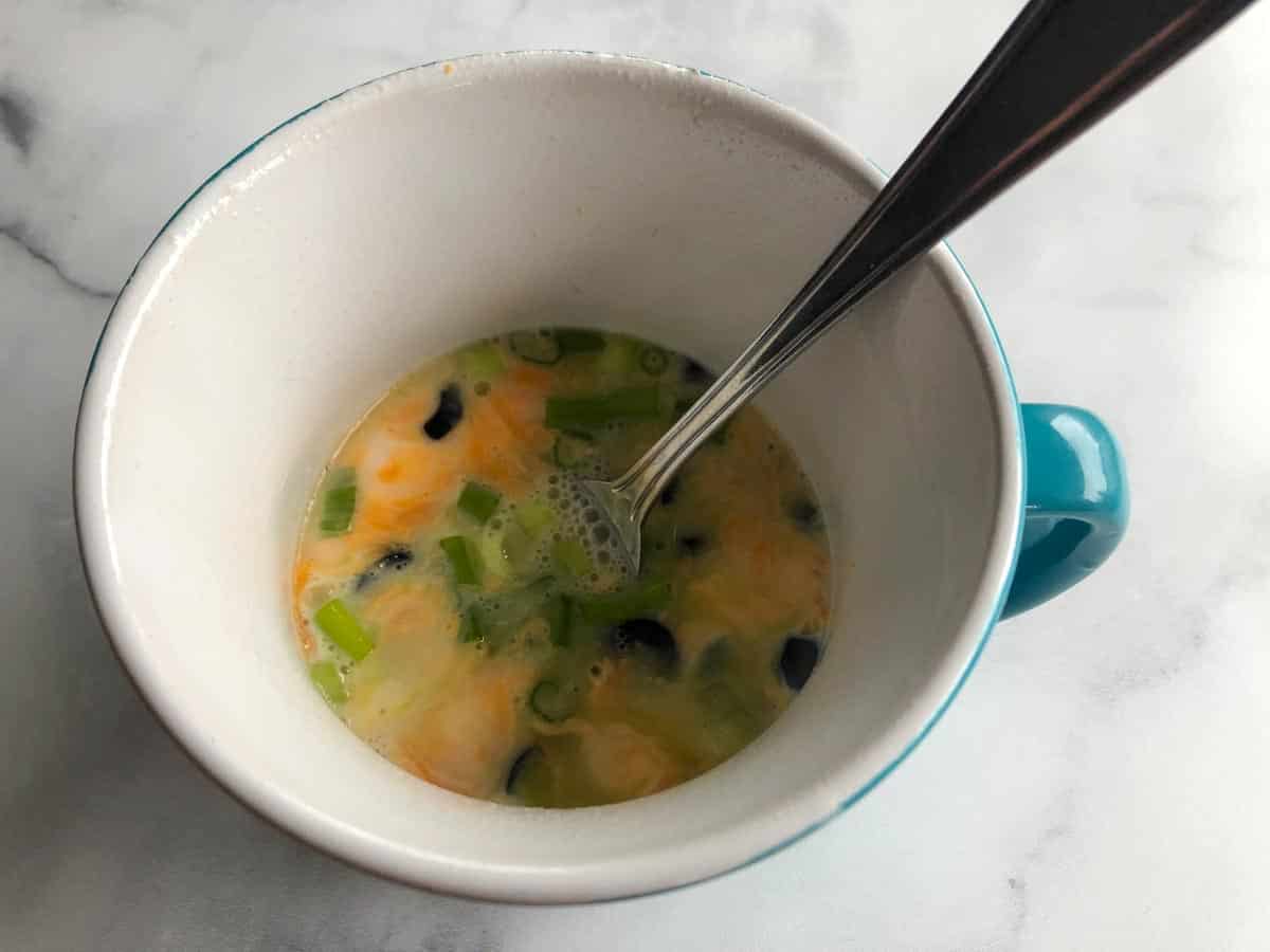 Omelette being beaten with a fork in microwave-safe mug.