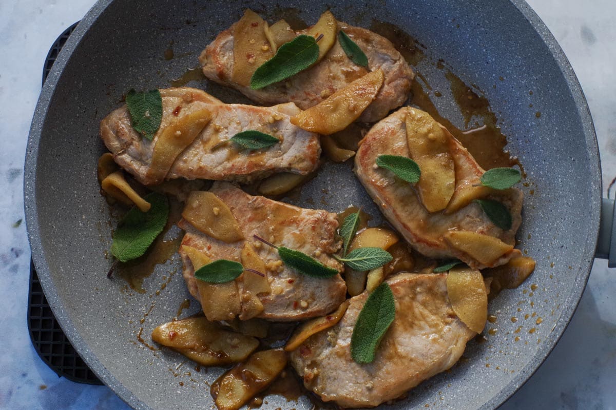 Cooked pork in pan with sauce and sage garnish.