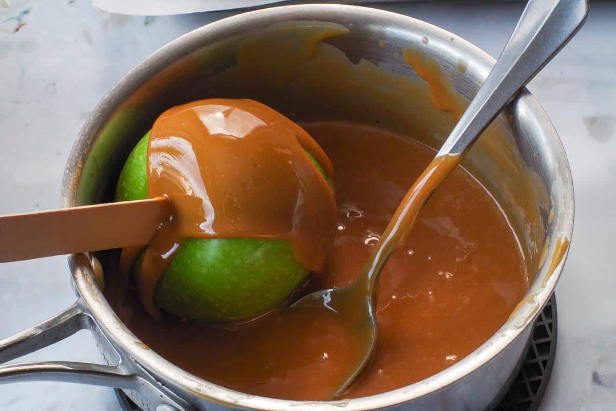 Apple being dipped in caramel and caramel being spooned over apple.