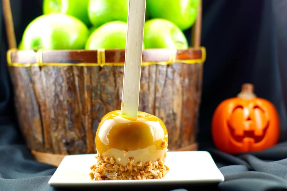 Salted Toffee Caramel Apple on a white plate with a basket of green apples and plastic jack o' lantern in the background.
