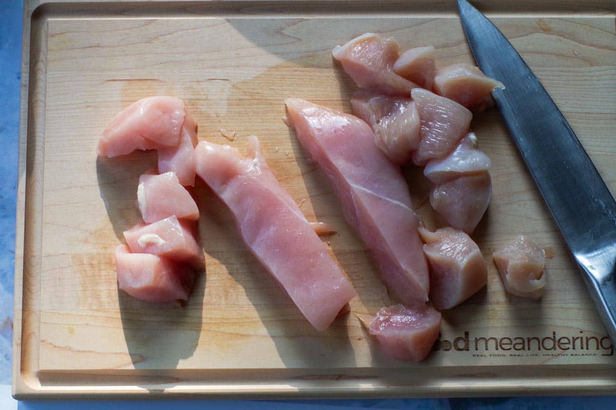 Chicken breast being cut into pieces on cutting board with large knife.