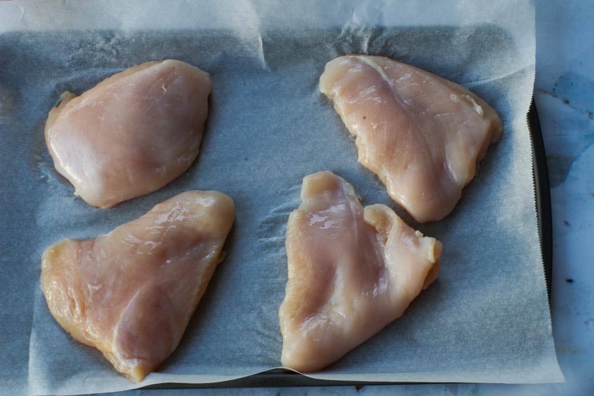 4 raw chicken breasts on a parchment-lined sheet pan.