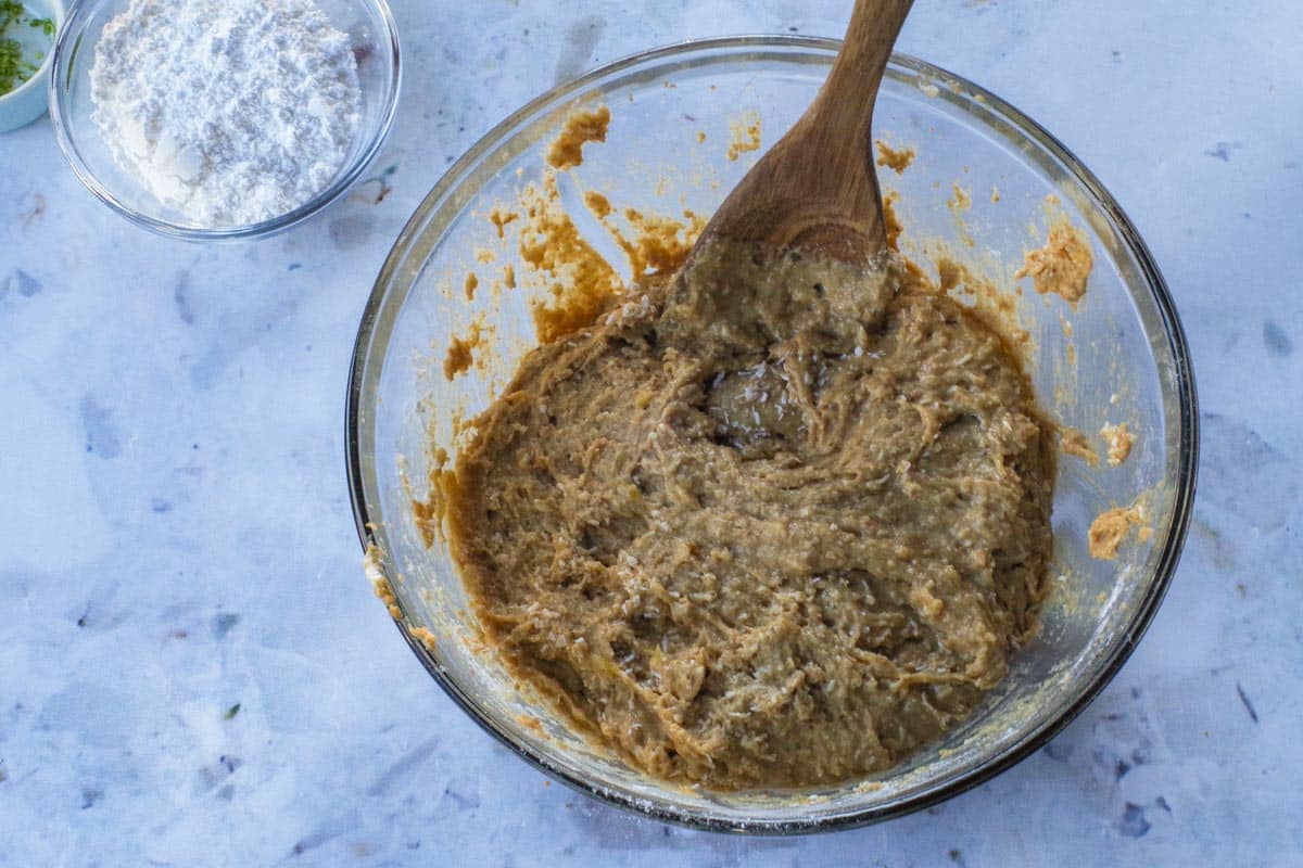 Coconut being stirred into batter with a wooden spoon, in a large glass bowl.