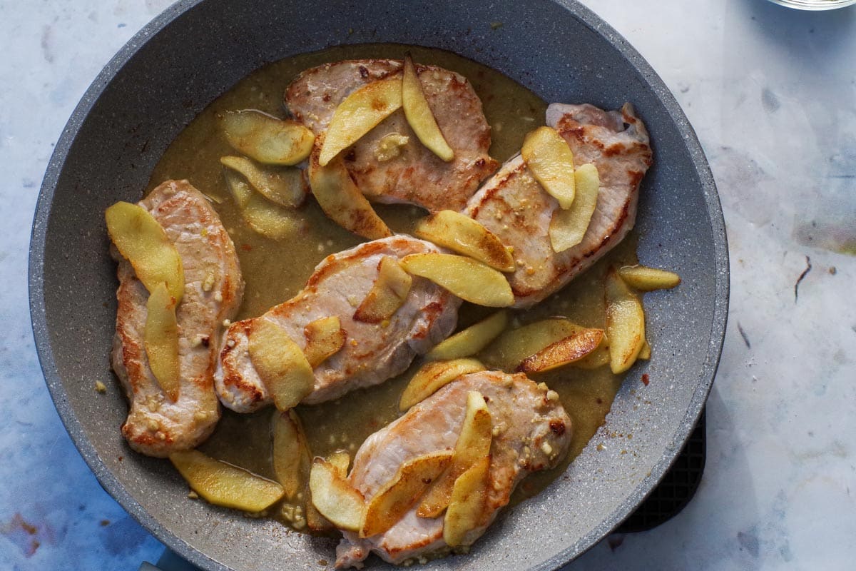 Pork with apples and sauce poured over it in frying pan.