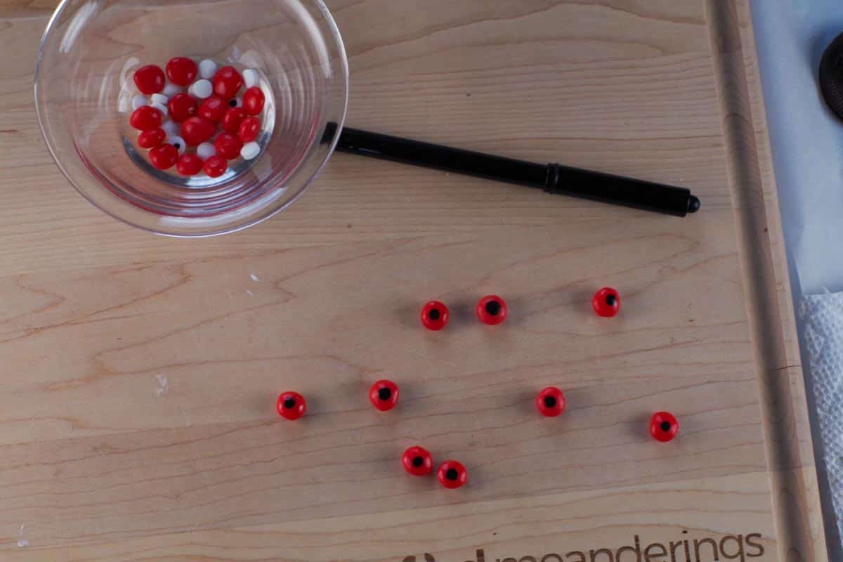 Red candy eyes on a cutting board with a black edible marker and more red candies and white candy eyes.