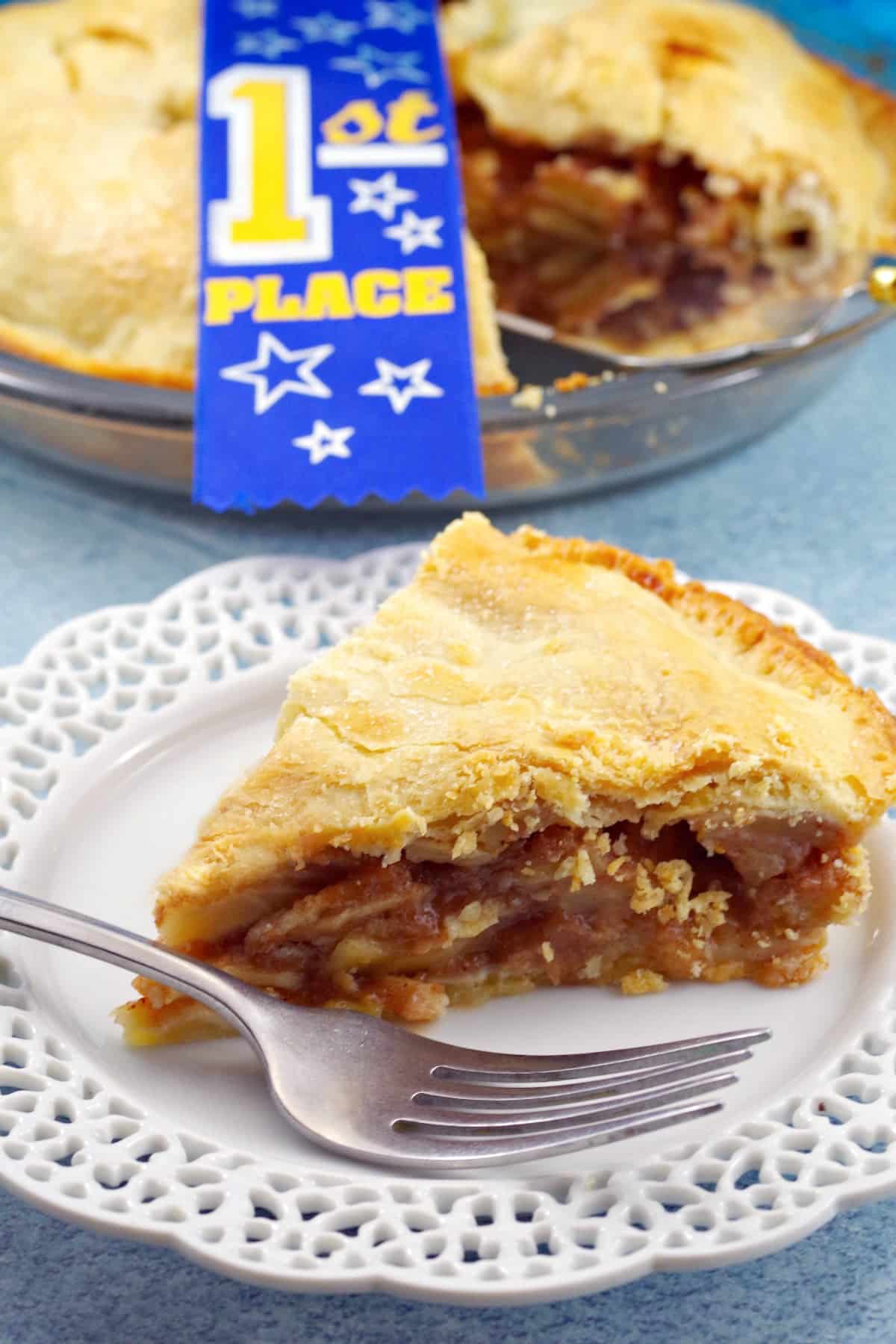 Slice of award-winning apple pie on a white plate with more pie in the background and a blue ribbon on the pie.
