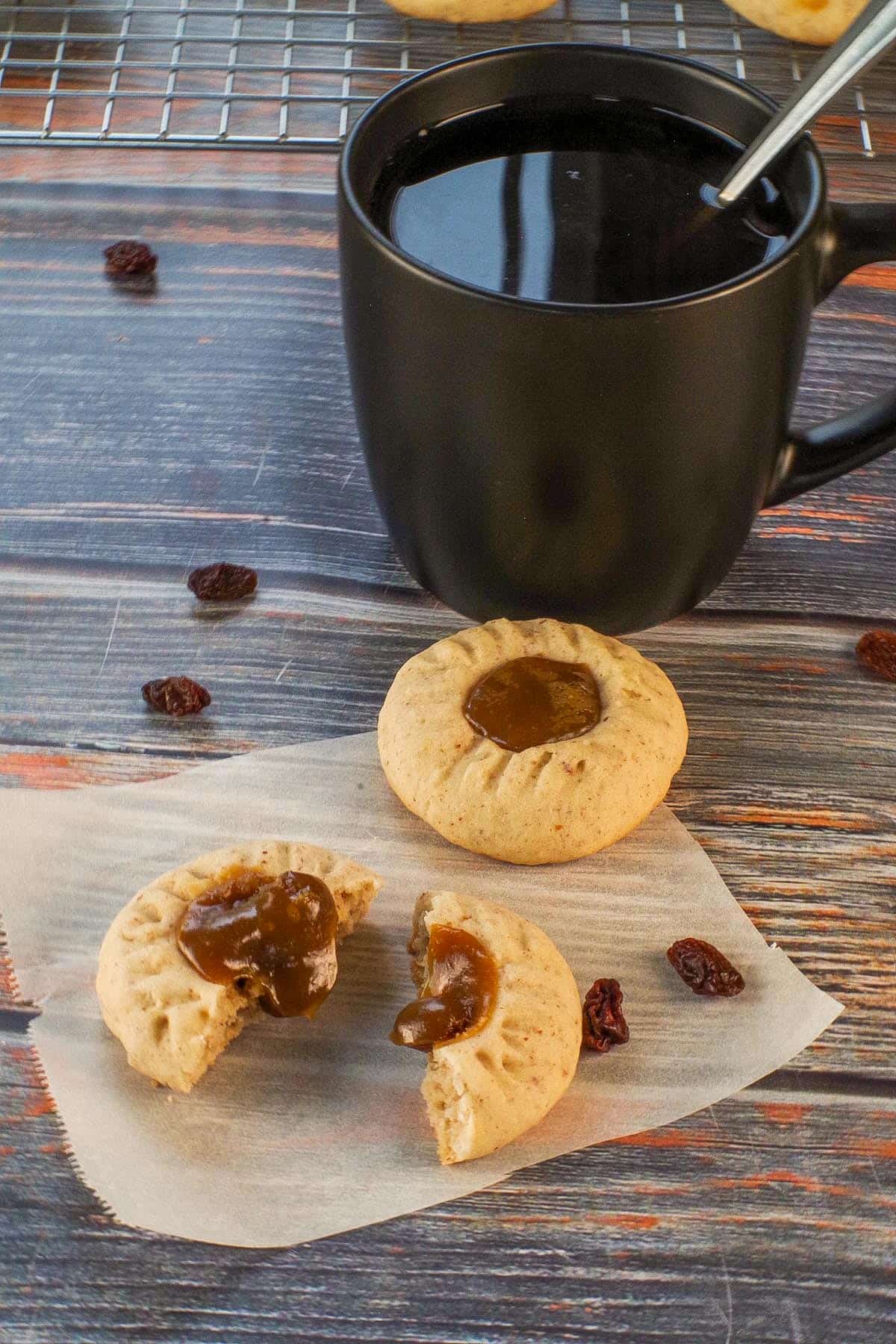 Butter tart cookies on parchment paper, with one split in half and filling oozing out. and black coffee mug in the background.