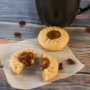 Butter tart cookies on parchment paper, with one split in half and filling oozing out. and black coffee mug in the background.