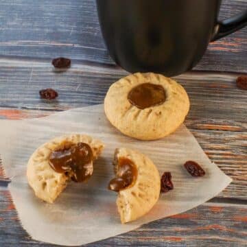 Butter tart cookies on parchment paper, with one split in half and filling oozing out. and black coffee mug in the background.
