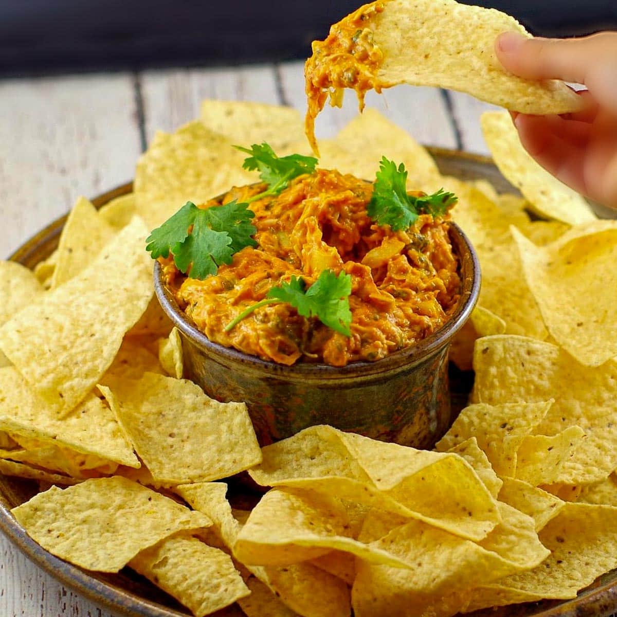 Baked artichoke pizza dip in the middle of a platter of tortilla chips with a hand dipping a chip into the dip bowl and lifting it out.