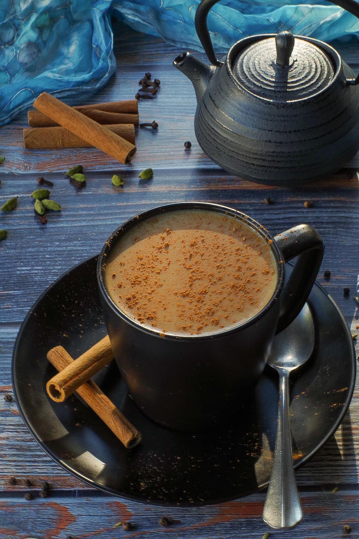 Masala Chai Tea in a black mug with a black tea pot, spices and a blue scarf in the background.