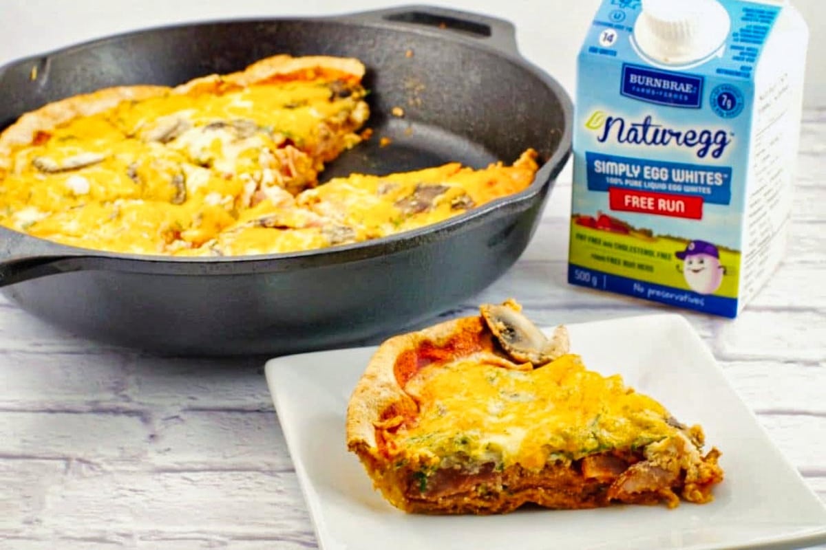A slice of healthy egg white pizza on a white plate with a carton of egg whites and a cast iron pan with more pizza, in the background.