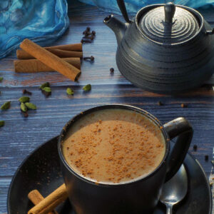 Masala Chai Tea in a black mug with a black tea pot, spices and a blue scarf in the background.