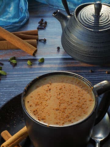 Masala Chai Tea in a black mug with a black tea pot, spices and a blue scarf in the background.