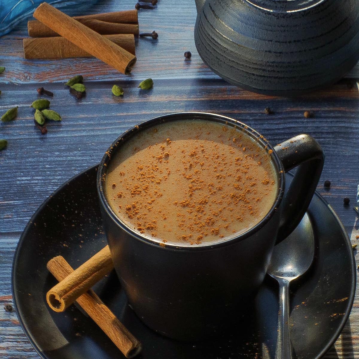 Masala Chai Tea in a black mug with a black tea pot, spices and a blue scarf in the background.