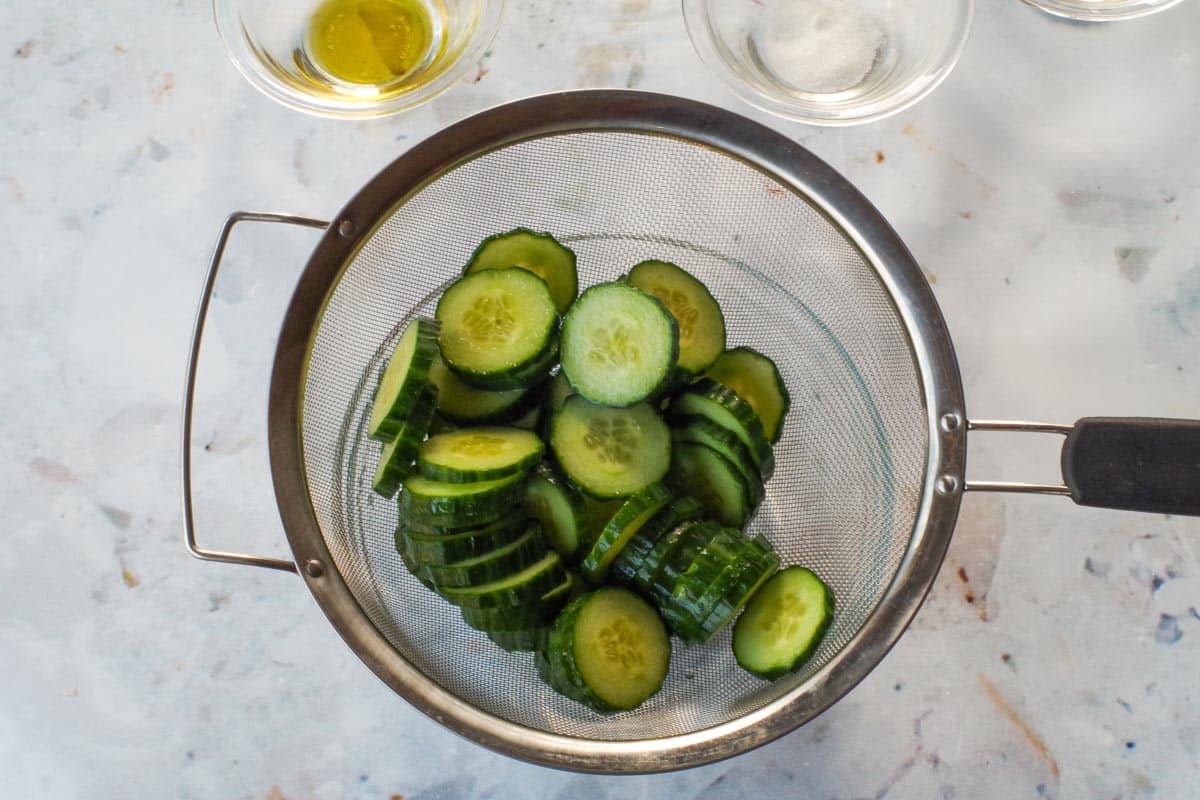 Cucmber slices being drained through a sieve over a glass bowl.