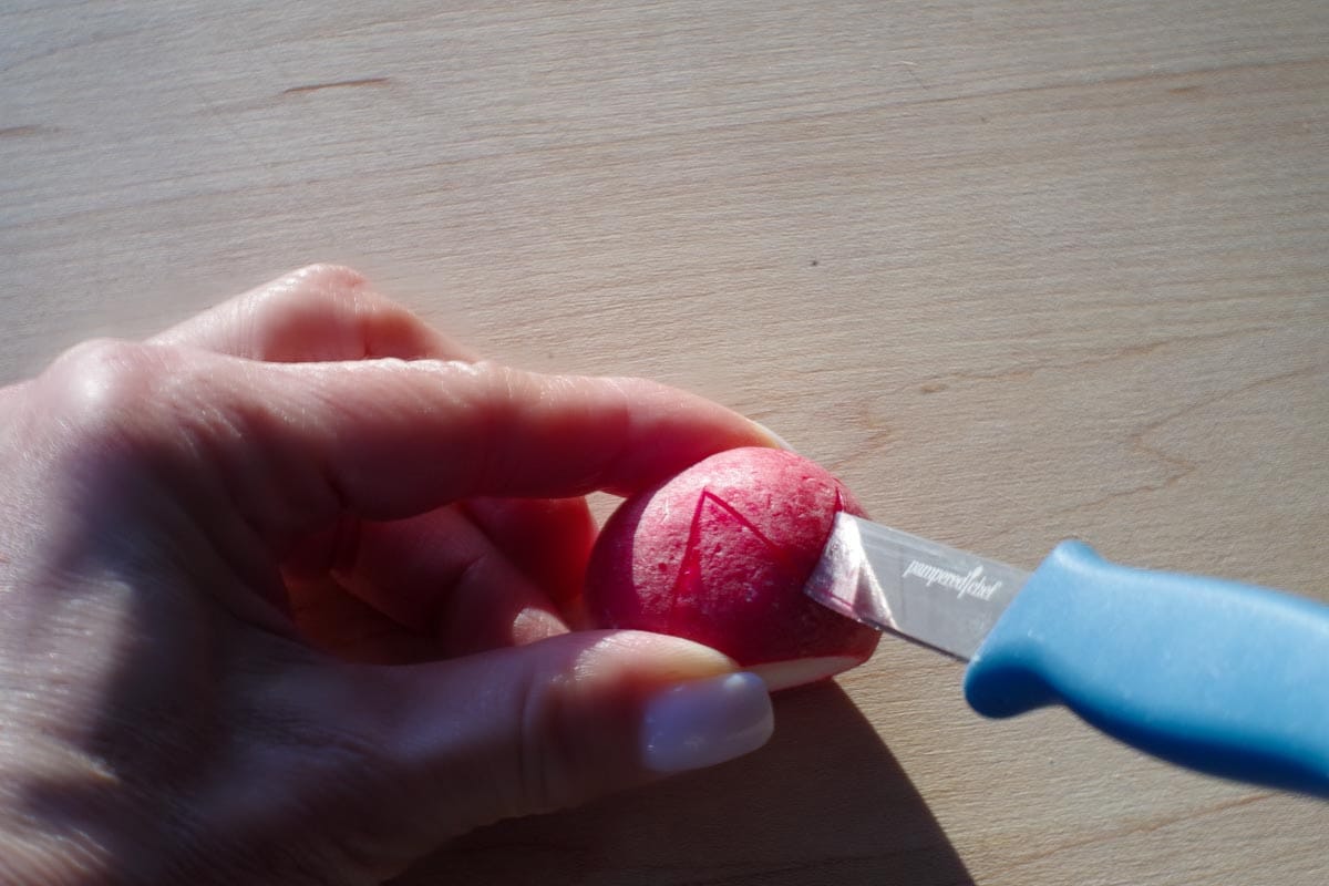 Zigzag pattern being cut into radish with a small paring knife on a wooden cutting board.