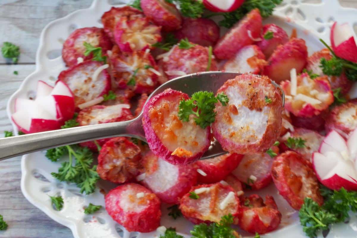 2 air fryer roasted radishes being held up on a spoon over a plate of roasted radishes.