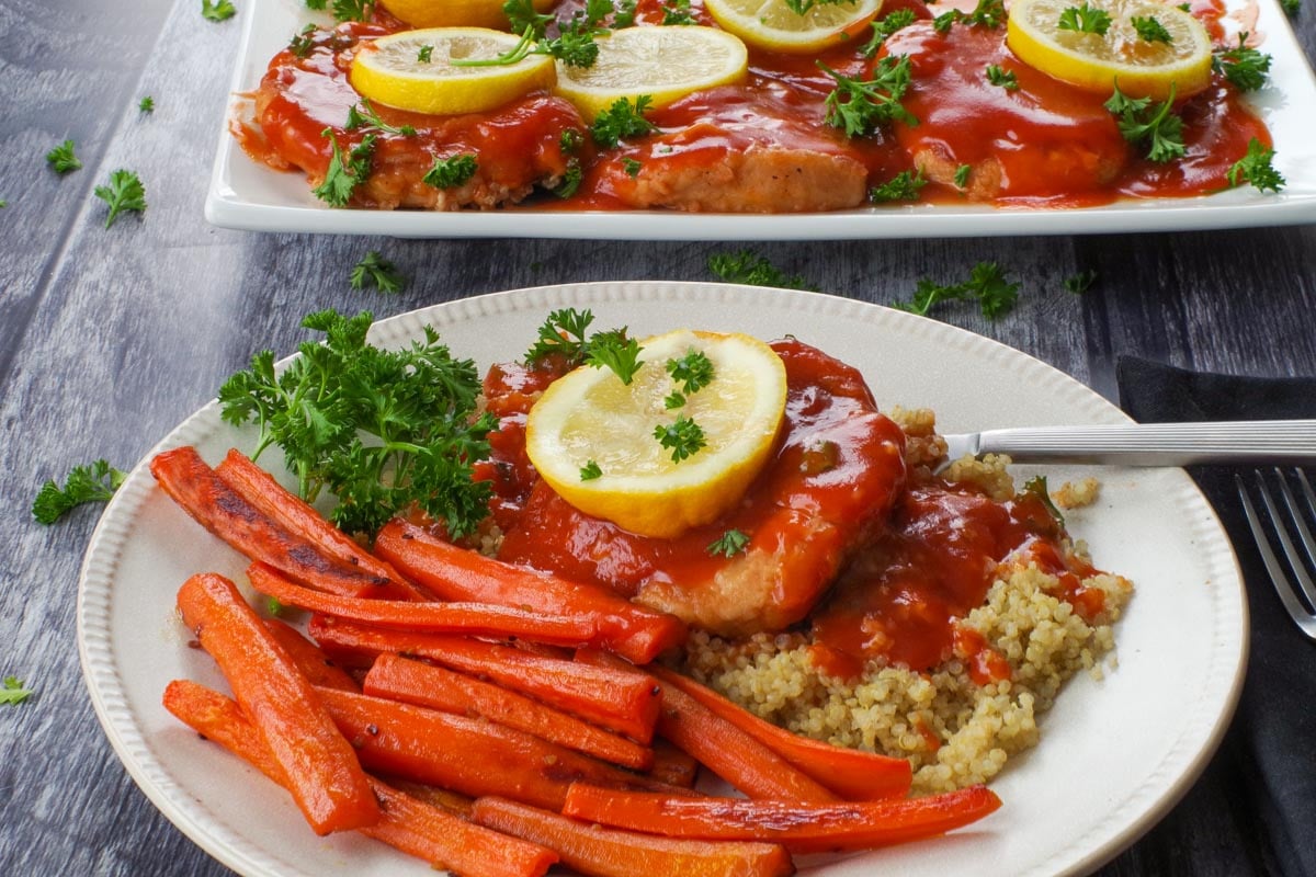 A baked pork loin chop with tangy sauce on a bed of quinoa, with a slice of lemon on top, roasted carrots on the side, and more pork on a platter in the background.