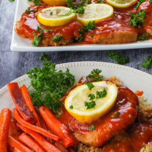 A baked pork loin chop with tangy sauce on a bed of quinoa, with a slice of lemon on top, roasted carrots on the side, and more pork on a platter in the background.