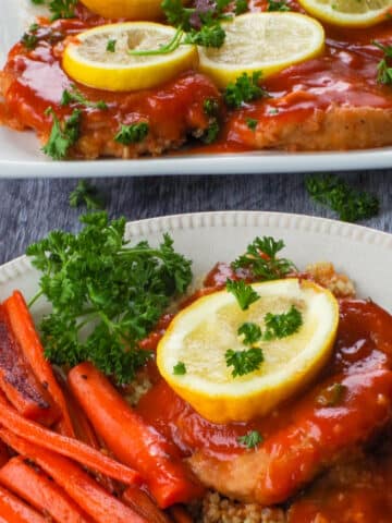 A baked pork loin chop with tangy sauce on a bed of quinoa, with a slice of lemon on top, roasted carrots on the side, and more pork on a platter in the background.