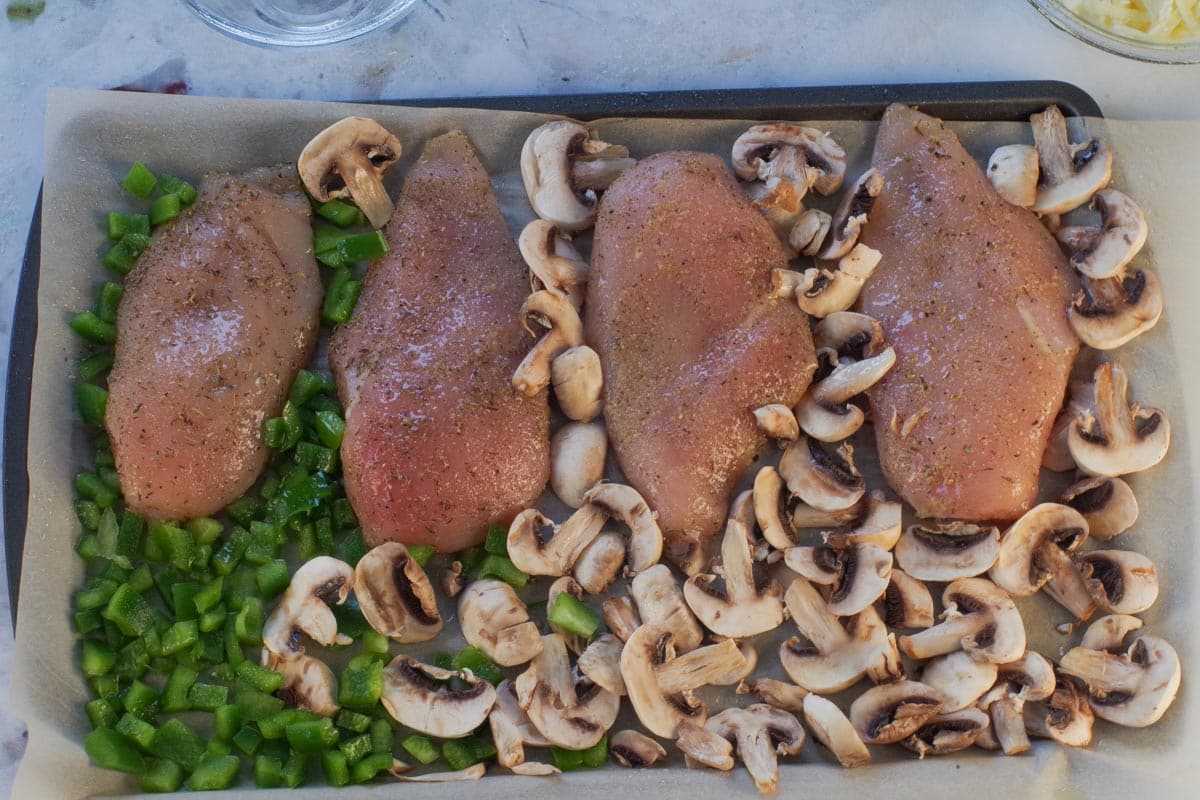 Raw rubbed chicken with uncooked chopped green peppers and sliced mushrooms on a sheet pan.