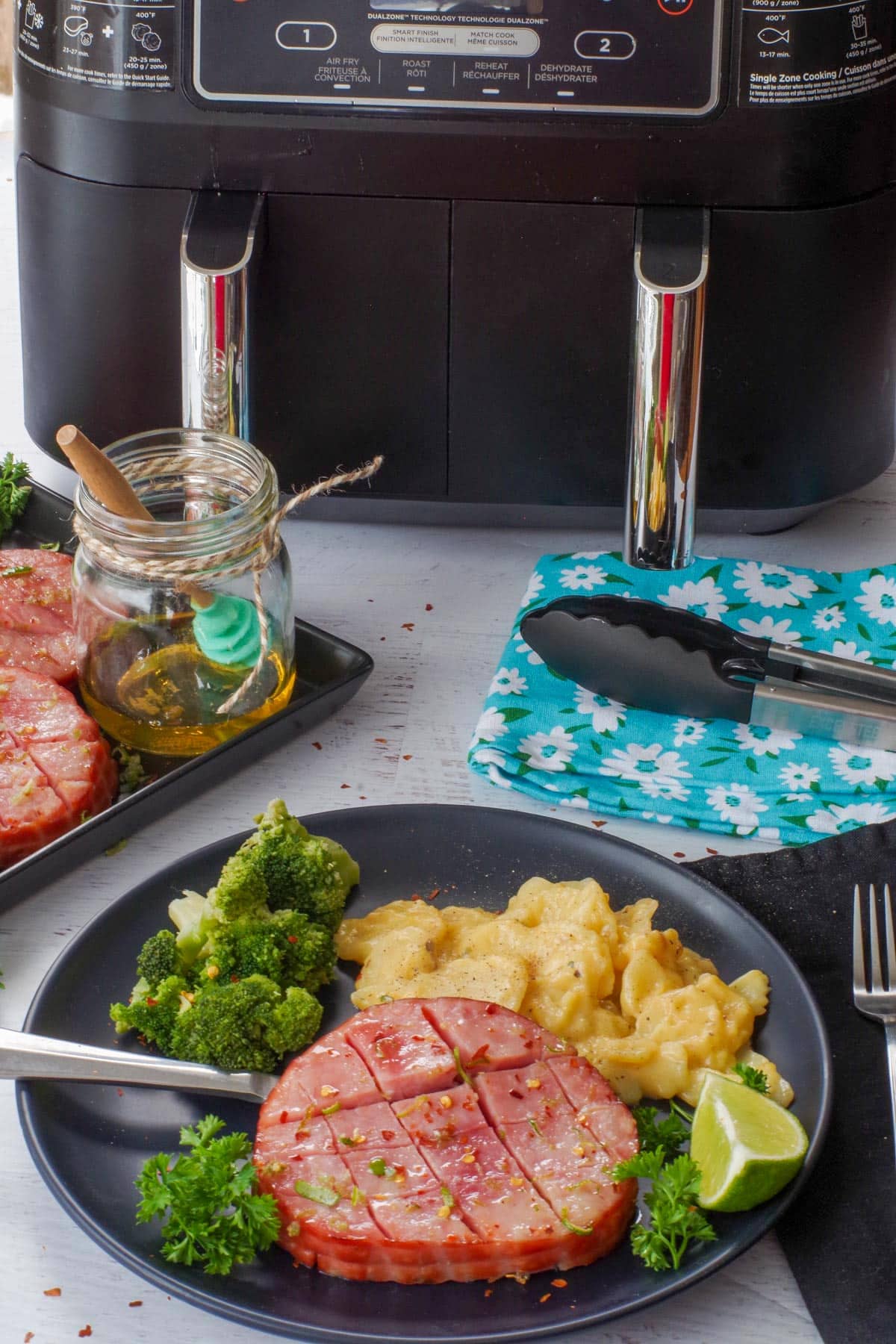 Air Fryer Ham Steak on a black plate with broccoli and scalloped potatoes, with a tray of more ham and a Ninja air fryer in the background.