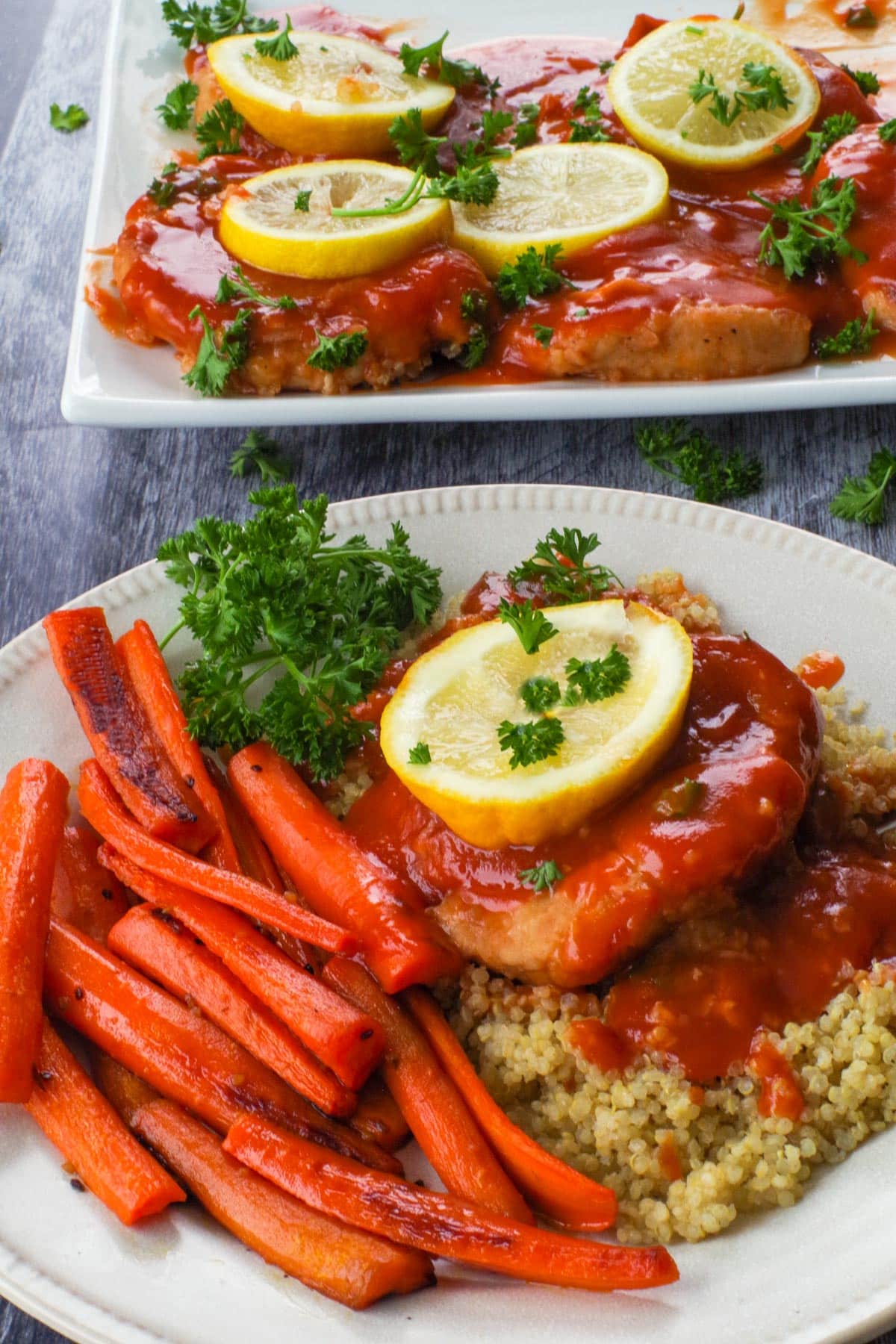 A baked pork loin chop with tangy sauce on a bed of quinoa, with a slice of lemon on top, roasted carrots on the side, and more pork on a platter in the background.