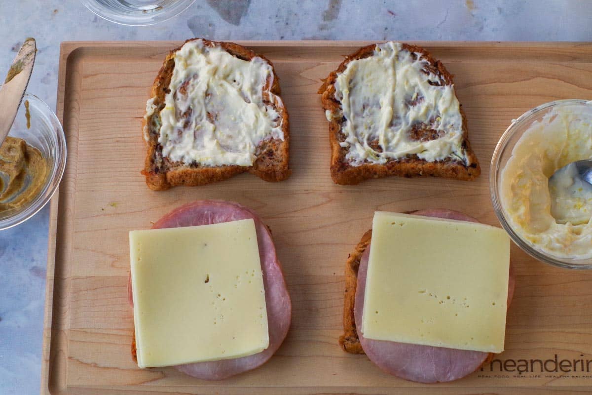 4 piece of coated bread on a wooden cutting board. 2 pieces have mayo lemon garlic mixture and other side has mustard, cheese and ham.