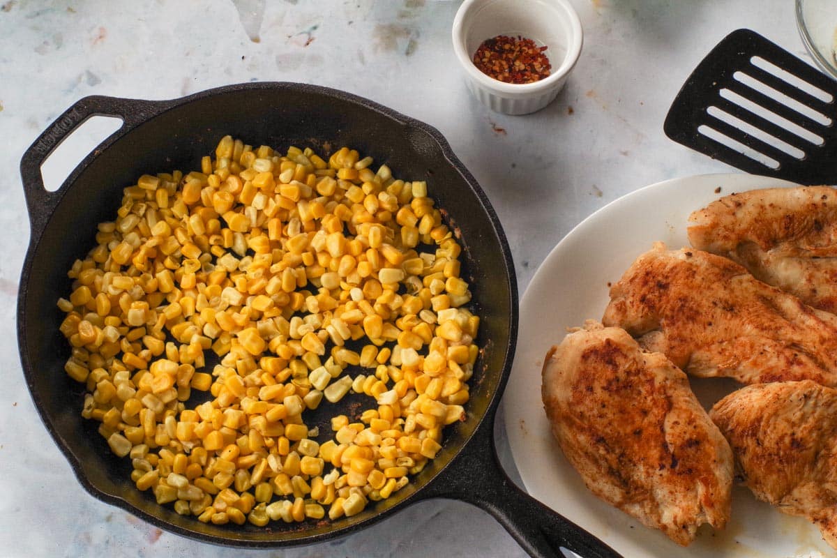 Corn in cast iron frying pan with chicken in a plate on the side.