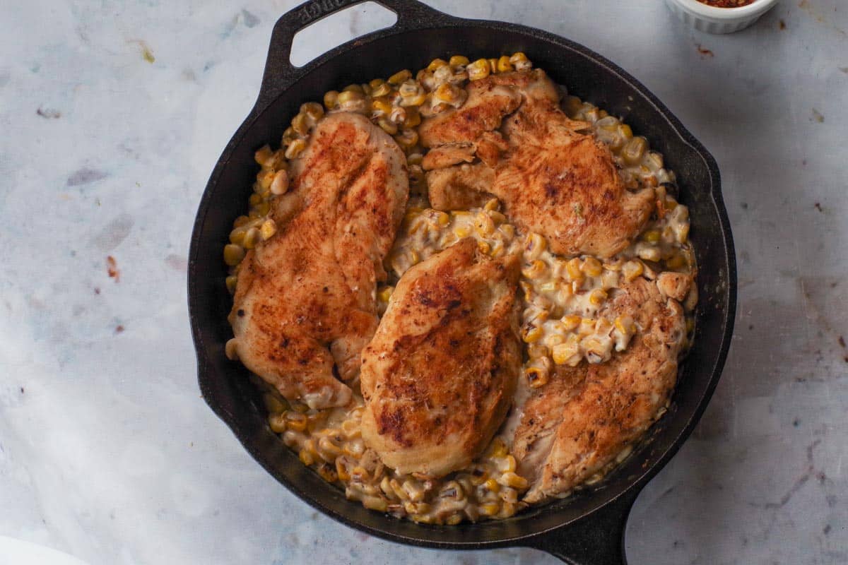 Chicken nestled into corn with sauce in a cast iron frying pan.