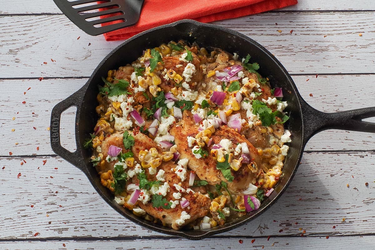Mexican Street Corn Chicken in a cast iron pan with an orange napkin and a black spatula in the background.