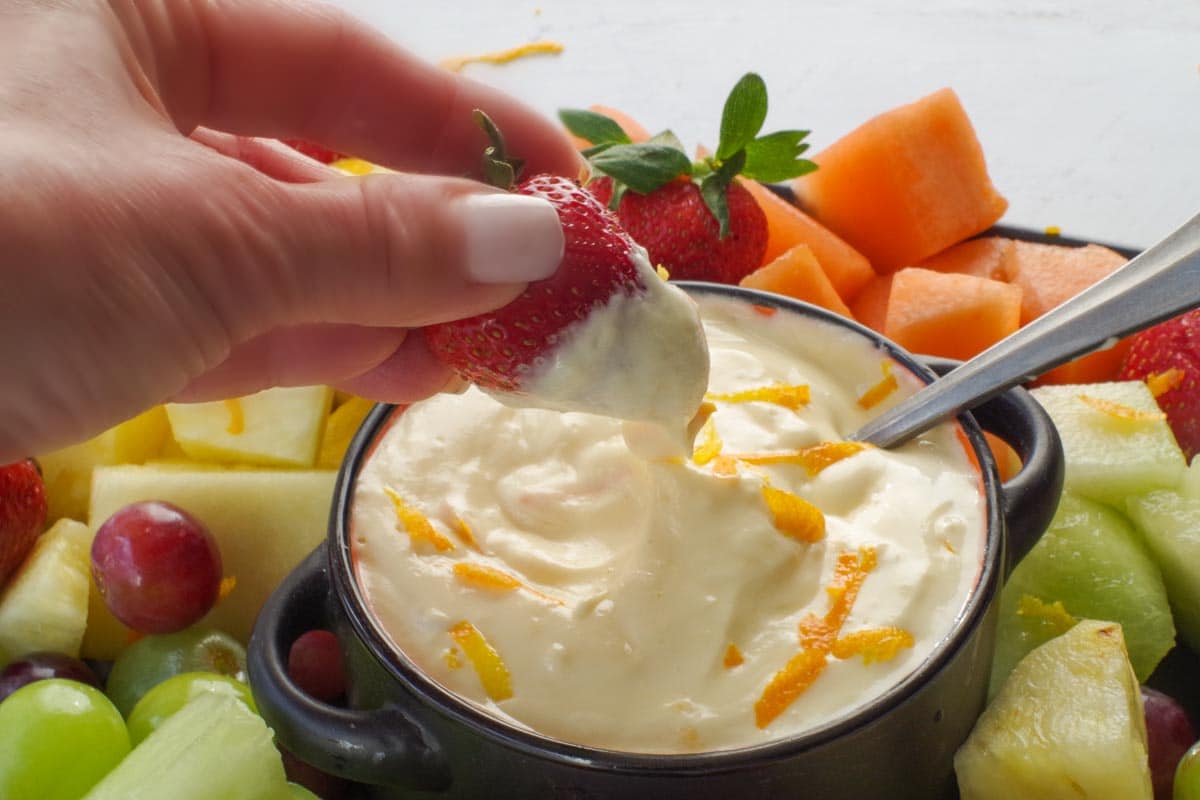 A strawberry being dipped into Orange Creamsicle Dip in a black bowl, surrounded by a variety of fruit, on a black tray.