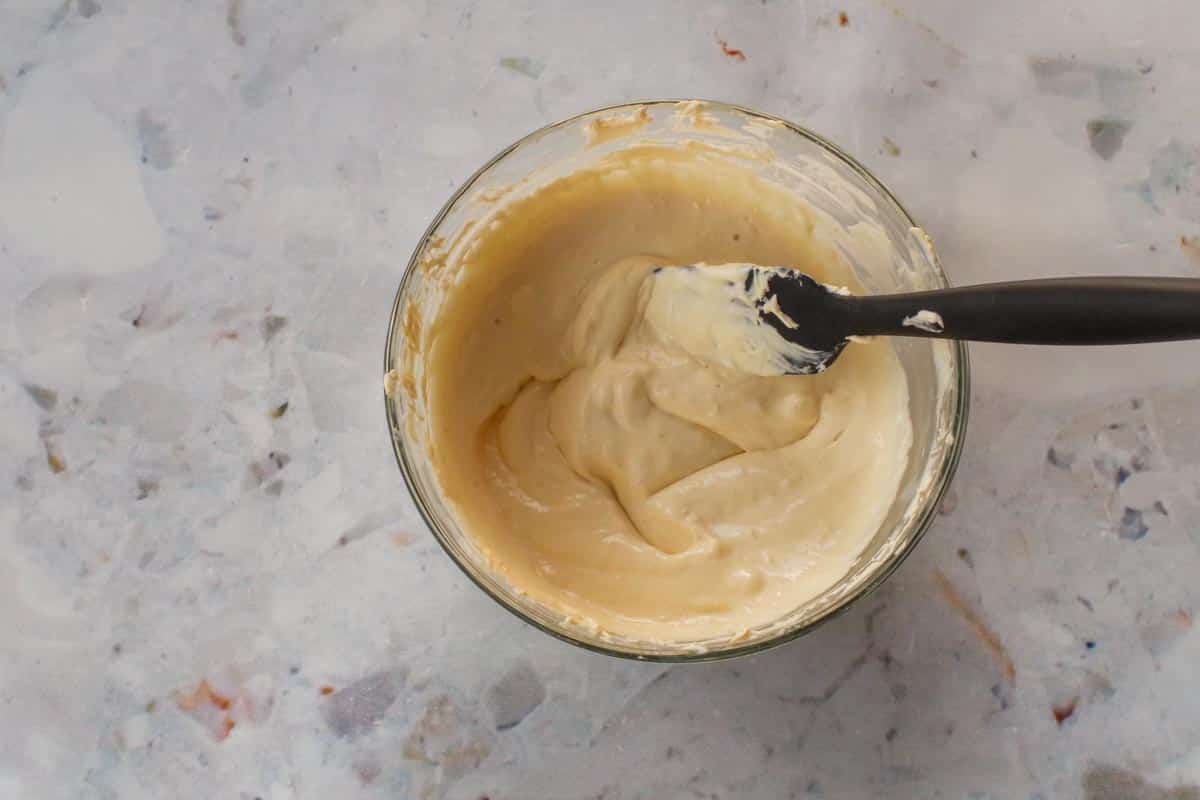 Marshmallow creme folded into orange cream cheese mixture in a glass bowl with a black silicone half spatula.