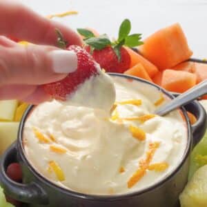 A strawberry being dipped into Orange Creamsicle Dip in a black bowl, surrounded by a variety of fruit, on a black tray.