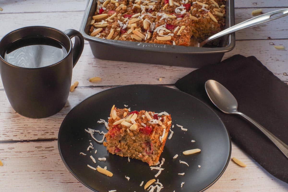 A slice of Baked Rhubarb Oatmeal on a black plate, with a cup of coffee and a pan with more Rhubarb Baked Oatmeal in the background.